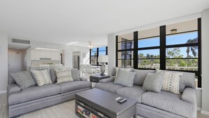 Un salon moderne avec un canapé gris, une table basse sur laquelle sont posés des livres et de grandes fenêtres donnant sur des palmiers.