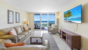 A living room with a sofa, armchair, coffee table, and TV. There is a balcony with a view of the ocean.