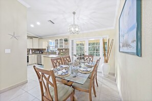 A dining area with a glass table, wooden chairs, and a chandelier. There is a kitchen in the background with cabinets and a microwave. - Pet-friendly Waterfront | Pool & Steps to Beach (Marco Island)