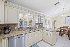 A kitchen with white cabinets, granite countertops, a dishwasher, and a dining area with wooden chairs and a table. - Pet-friendly Waterfront | Pool & Steps to Beach (Marco Island)