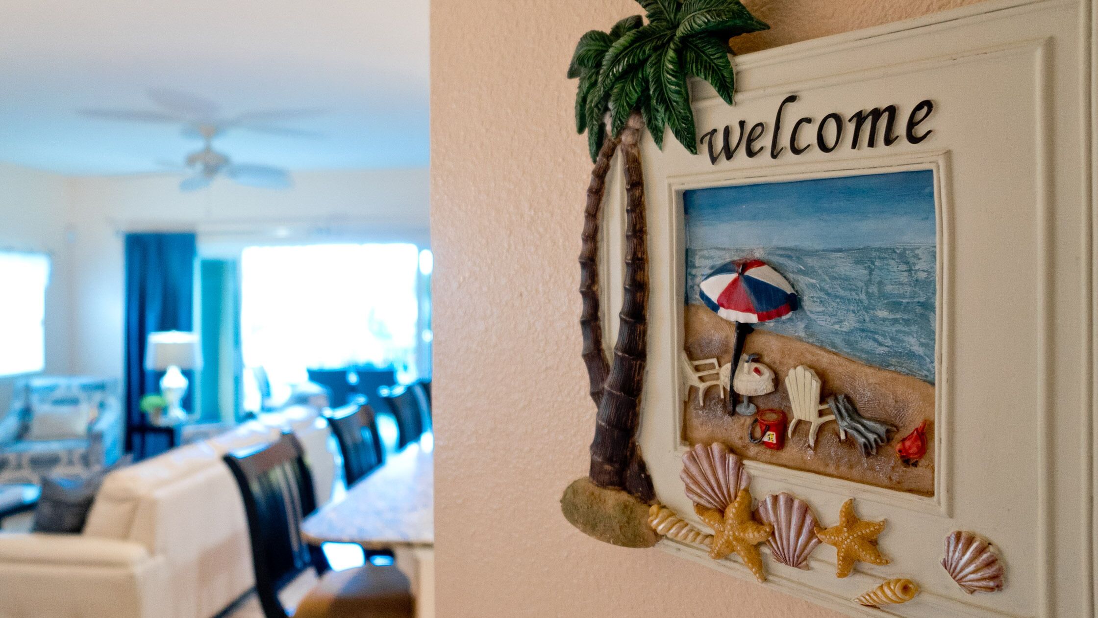 A living room with a framed welcome sign, a beach-themed painting, and a ceiling fan.