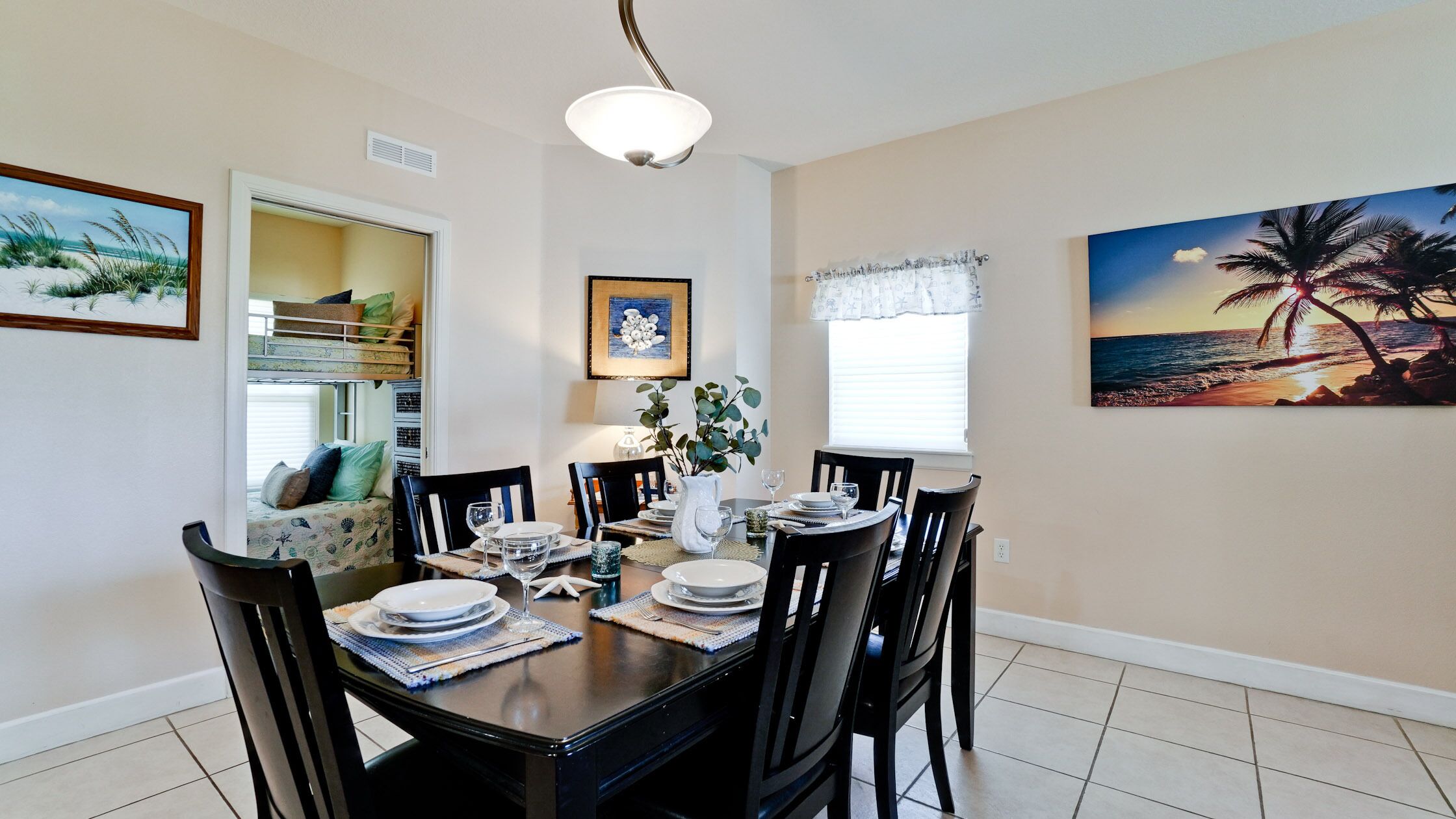 A dining area with a wooden table set for four, surrounded by chairs. There are framed artworks on the walls, a window with blinds, and a ceiling light fixture.