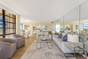 A spacious living area with a large window, a dining table, and a kitchen in the background.
