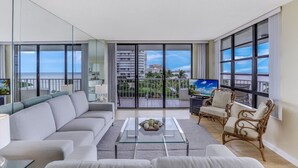 A modern living room with a large window, a sofa, a coffee table, and wicker chairs. - Front Unit Facing Beach | Wraparound Balcony (Marco Island)