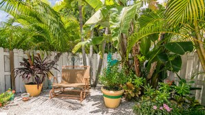 A tropical garden with a wooden chair, potted plants, and a white fence. - Sail Away Cottage · 170 Steps · Pool · 1920's Cottage (Bradenton Beach)