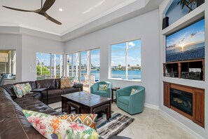 A spacious living room with a large sectional sofa, teal armchairs, and a wooden coffee table. A fireplace and a large window with a view of the ocean are also visible.
