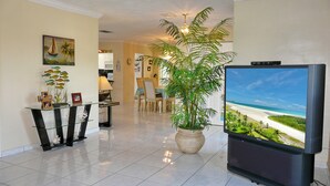 Amplia sala de estar con un televisor de pantalla plana grande, una mesa de comedor, sillas, una planta en maceta y una cocina al fondo.
