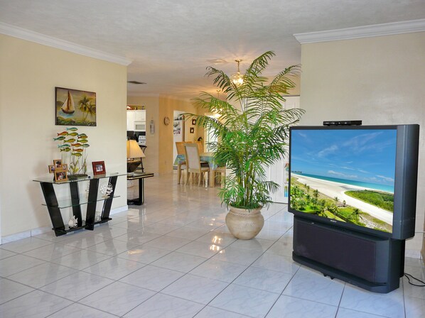A spacious living area with a large flat-screen TV, a dining table, chairs, a potted plant, and a kitchen in the background.
