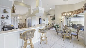 Una cocina amplia con techos altos, armarios blancos y una isla central. Hay taburetes de mimbre y un comedor con candelabro.