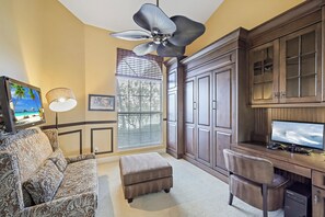A room with a large wooden desk, a computer, a television, a patterned sofa, and a ceiling fan.