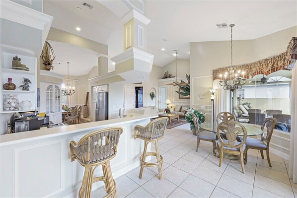 A spacious kitchen with high ceilings, white cabinetry, and a central island. There are wicker bar stools and a dining area with a chandelier.