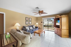 A living room with a sofa, coffee table, and TV stand. There are two lamps, a ceiling fan, and a sliding glass door leading to an outdoor area. - Boater’s Paradise | Unmatched Water Views (Marco Island)
