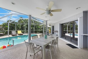 A poolside area with a table and chairs, a ceiling fan, and a view of the pool and palm trees.