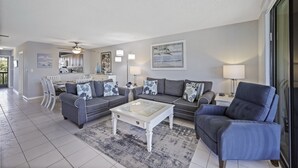 A spacious living room with a dining area, grey sofas with blue floral cushions, a glass coffee table, and a ceiling fan. - South End Condo | Pool, Spa and Steps to Beach (Marco Island)