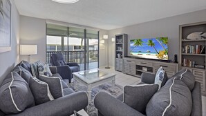 A modern living room with a large flat-screen TV, grey sofas, a glass coffee table, and a bookshelf. - South End Condo | Pool, Spa and Steps to Beach (Marco Island)