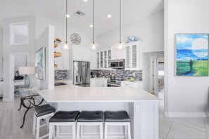 Una cocina moderna con gabinetes blancos, una isla grande con taburetes de bar, electrodomésticos de acero inoxidable e iluminación colgante.