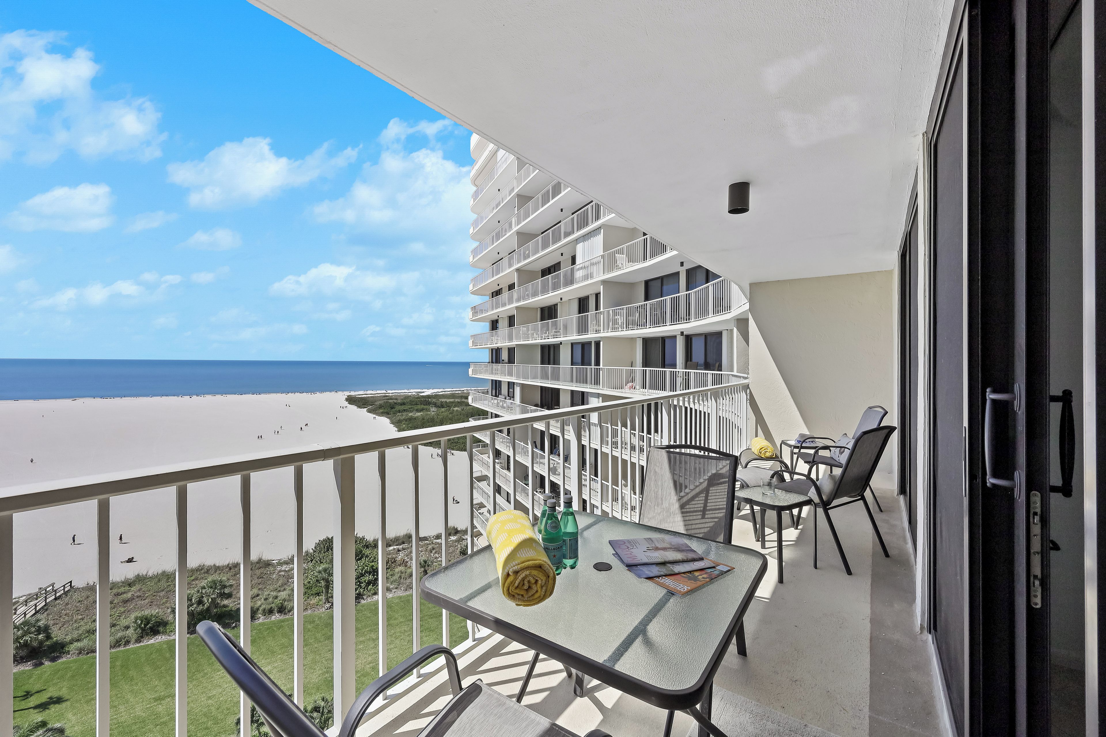 A balcony with a glass table, chairs, and a view of the beach.