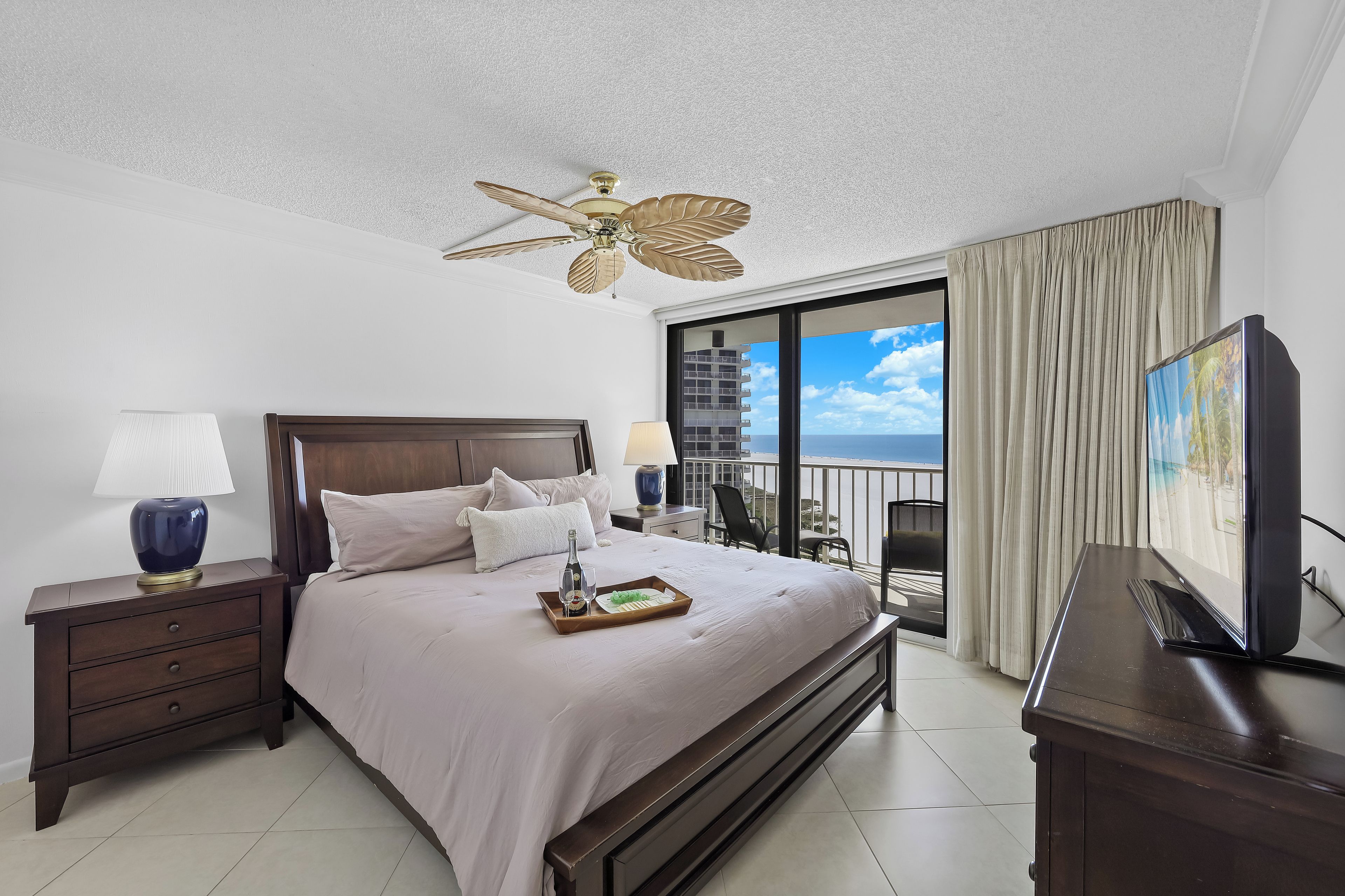Habitación de hotel con una cama grande, ventilador de techo, televisor, mesita de noche y balcón con vista al océano.