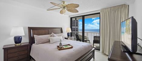 Habitación de hotel con una cama grande, ventilador de techo, televisor, mesita de noche y balcón con vista al océano.