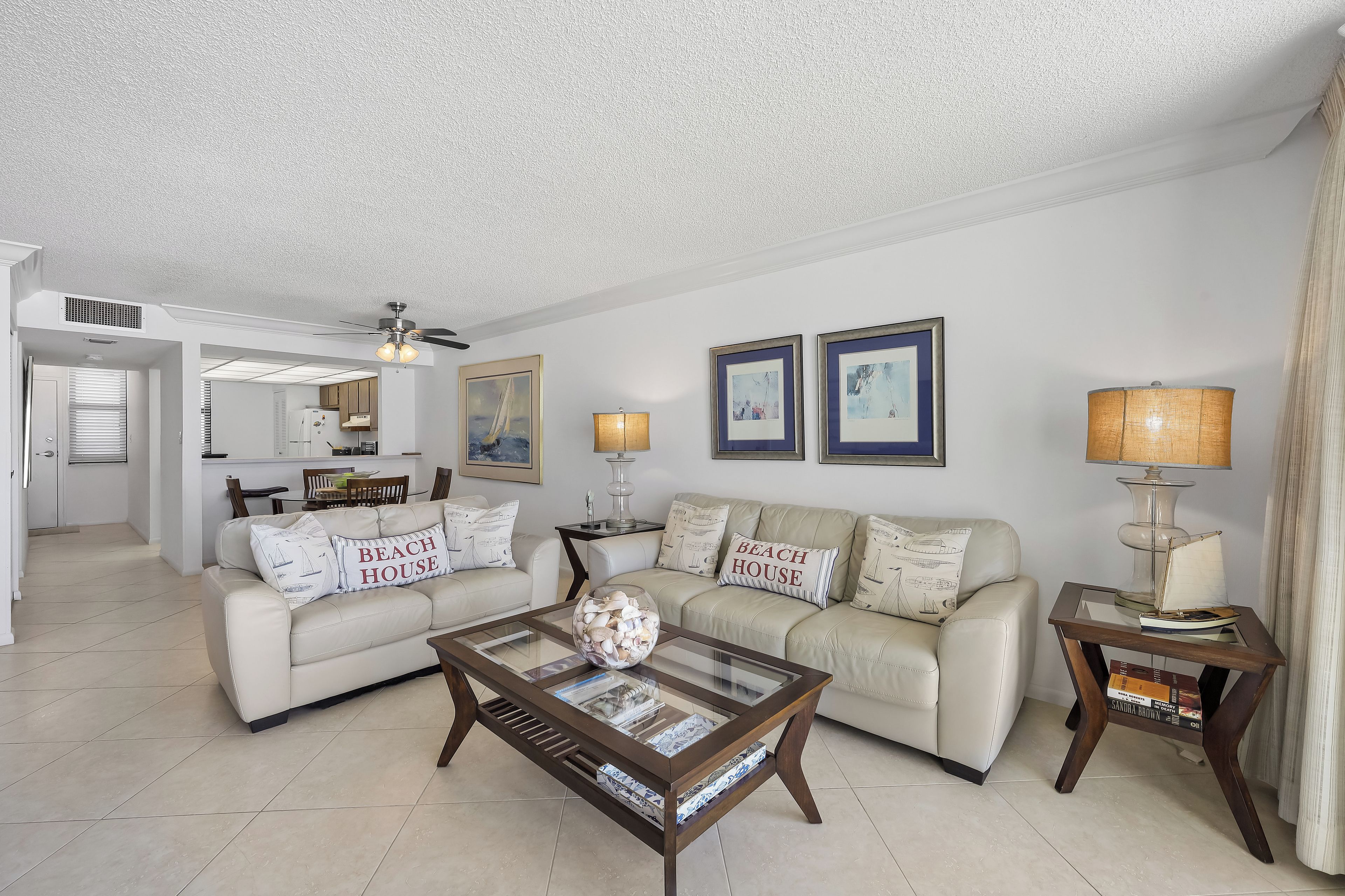 A living room with a sofa, coffee table, and a lamp. There is a kitchen in the background.