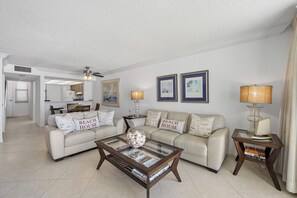 A living room with a sofa, coffee table, and a lamp. There is a kitchen in the background.