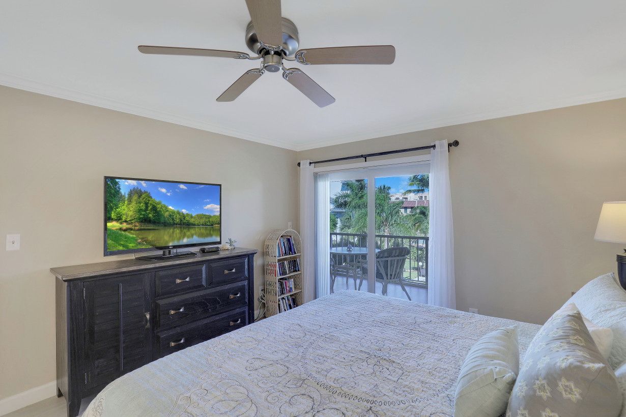 Une chambre à coucher comprenant un lit, une télévision, un ventilateur de plafond, une fenêtre avec vue et un balcon.