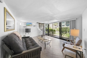A modern living room with a sofa, a coffee table, and a lamp. There is a large window with a view of greenery and buildings outside.