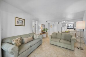 A spacious living area with two sofas, a small table with a lamp, and a kitchen in the background.