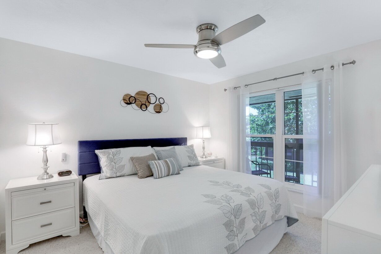 Un dormitorio con cama, mesitas de noche, ventilador de techo y ventana con cortinas.