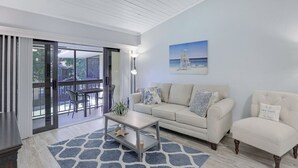 A modern living room with a sofa, armchair, and coffee table. There is a painting on the wall and a view of the outdoors through large sliding glass doors. - Charming Condo | Two Pools and Steps to Beach (Marco Island)