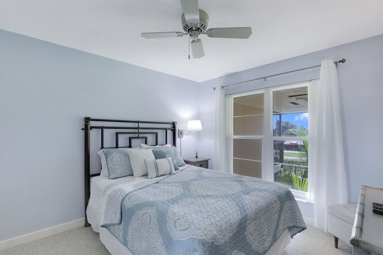A bedroom with a bed, a ceiling fan, a nightstand with a lamp, a window with curtains, and a view of greenery outside.