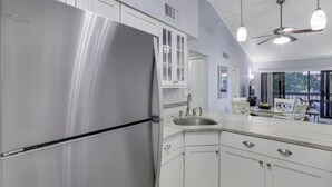 A modern kitchen with white cabinetry, a stainless steel refrigerator, and a dining area with a table and chairs. - Charming Condo | Two Pools and Steps to Beach (Marco Island)