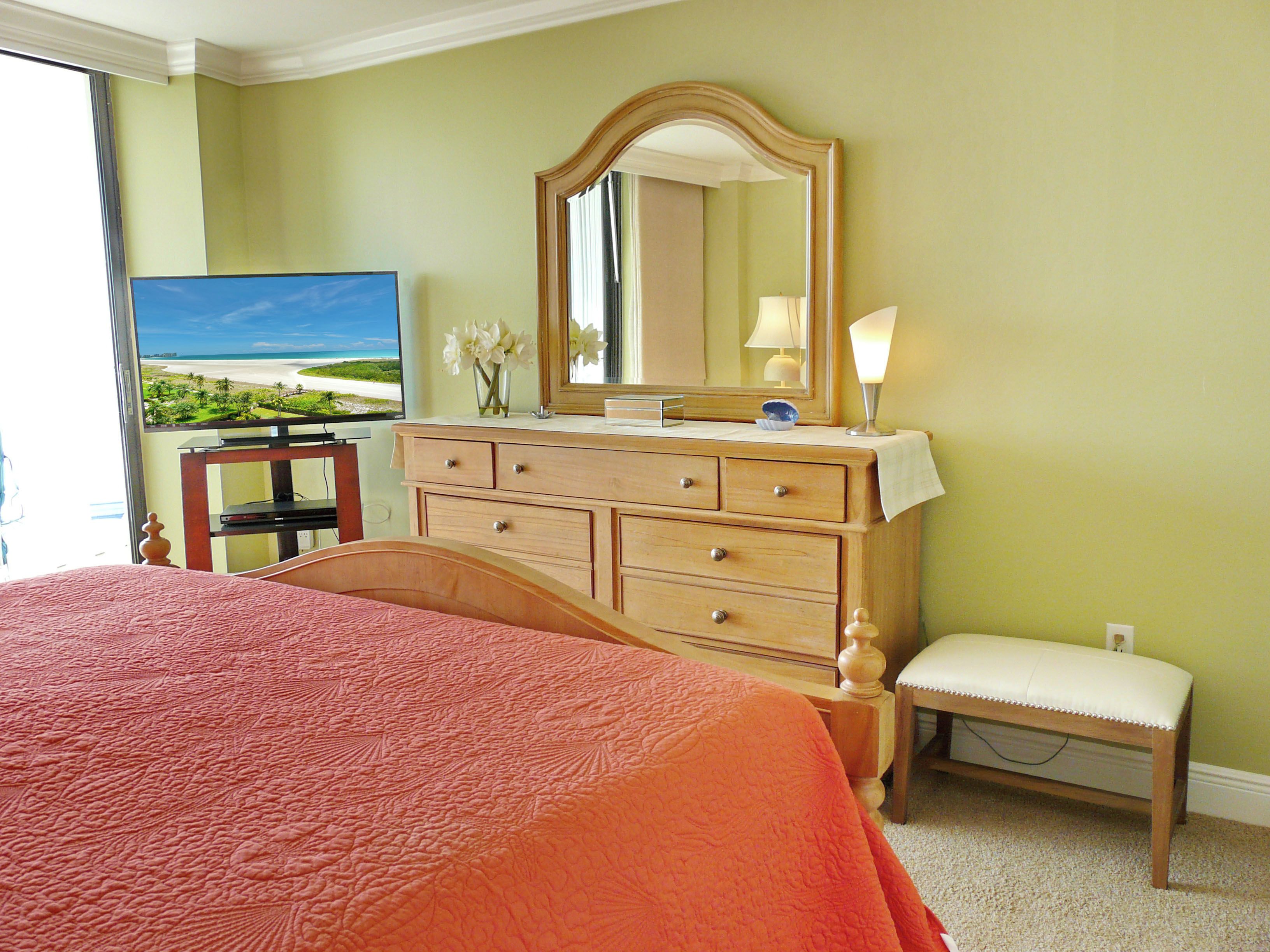 Een slaapkamer met een groot bed, een dressoir met spiegel, een televisie en uitzicht op een strand door het raam.