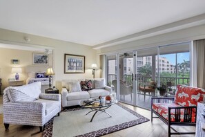 A spacious living room with a sofa, armchairs, and a coffee table. There is a balcony with a view of buildings and greenery.