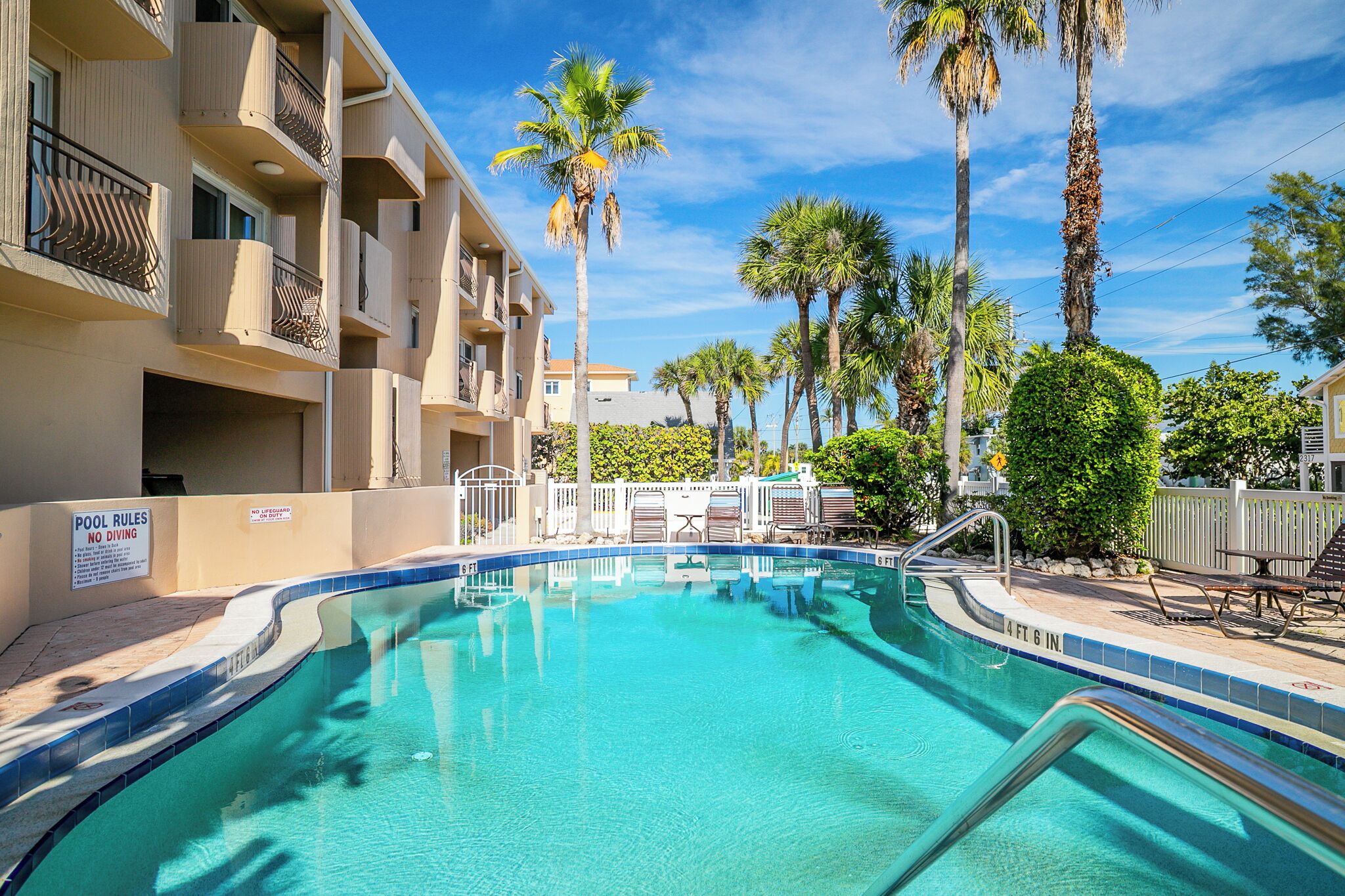 A swimming pool with a sign indicating no diving, surrounded by palm trees and a multi-story building with balconies.