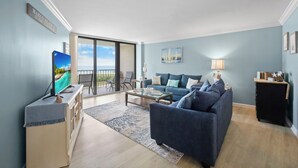 Ein Wohnzimmer mit einer Couch, einem Couchtisch und einem Fernseher auf einem Schrank. Eine Glastür führt auf einen Balkon.