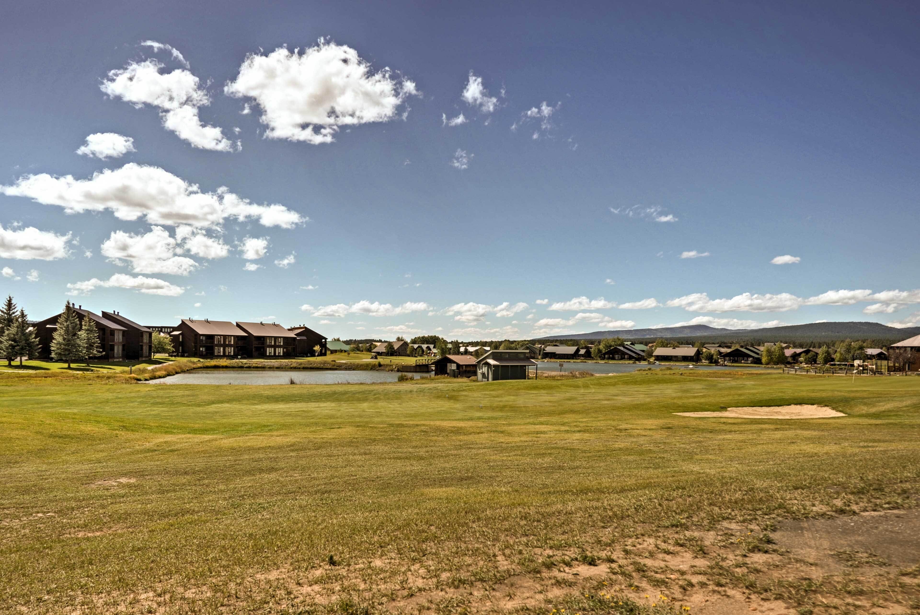 Un campo de golf con un estanque, edificios y montañas al fondo.