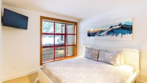 A bedroom with a bed, a window with a view, a painting on the wall, and a television mounted on the wall. - Cozy Retreat in The Woods, Hot Tub & Free Parking (Whistler)