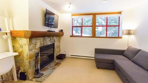 A living room with a stone fireplace, a grey sofa, a television mounted above the fireplace, and a window with a view of trees. - Cozy Retreat in The Woods, Hot Tub & Free Parking (Whistler)