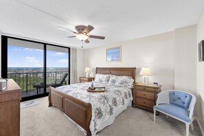 A bedroom with a large bed, a ceiling fan, a nightstand with lamps, a chair, and a view of the city through a balcony. - Beachfront Condo | Pool & Ocean View From Balcony (Marco Island)