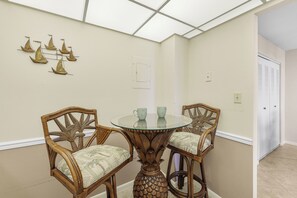 A dining area with a round glass table and four wicker chairs, a ceiling light fixture, and a door with louvered shutters. - Beachfront Condo | Pool & Ocean View From Balcony (Marco Island)
