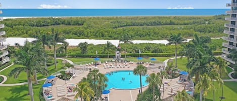 A resort with a central pool surrounded by palm trees and lounge chairs, overlooking a beach and ocean.