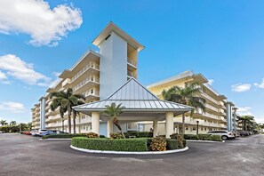 A hotel with a central atrium, multiple balconies, and a parking area with cars. - Coastal Style Condo | Pool, Spa & Steps to Beach (Marco Island)