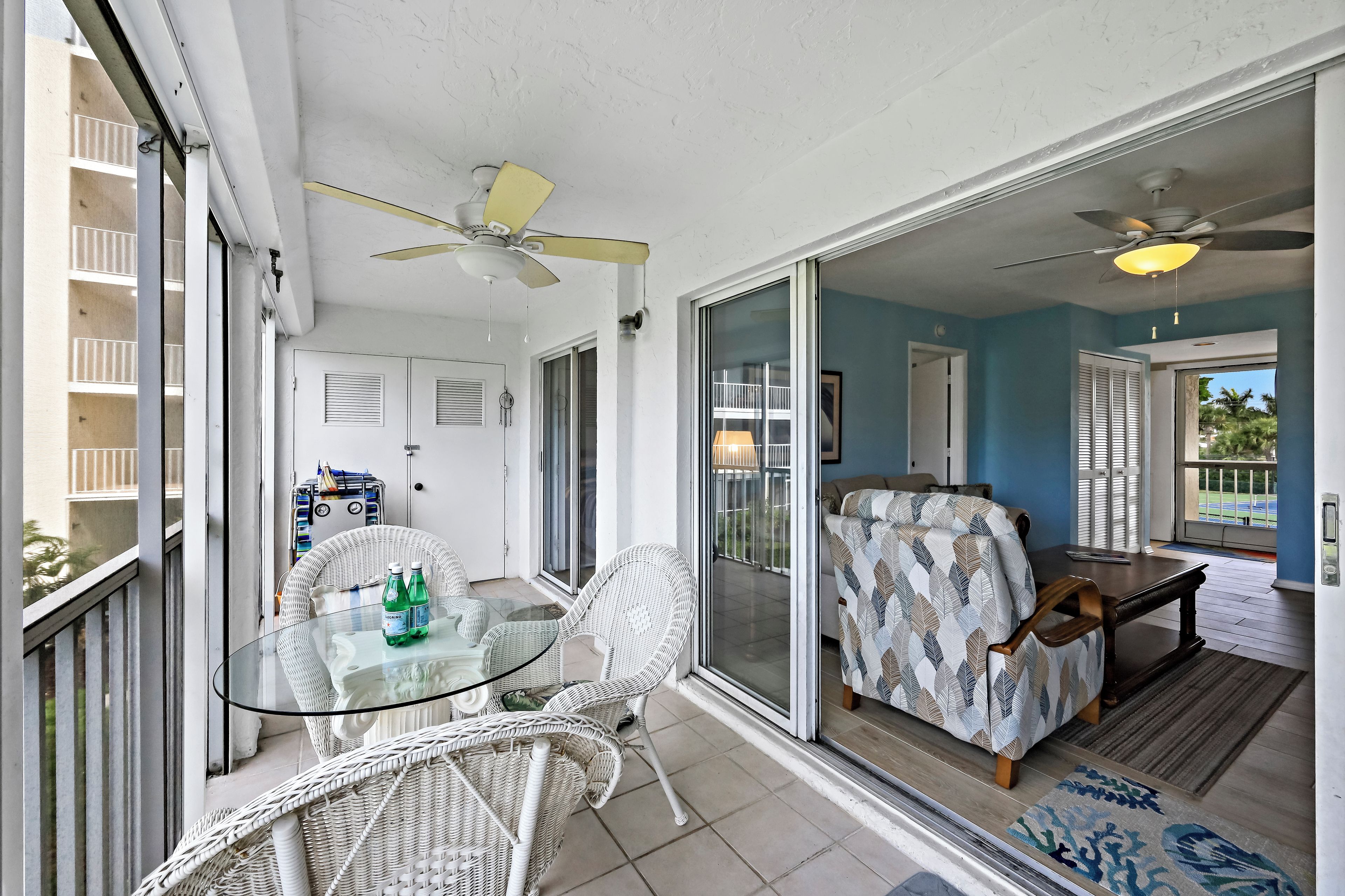 A balcony with a glass table, wicker chairs, ceiling fans, and a view of the outside.
