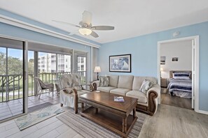 A living room with a sofa, chairs, a coffee table, a ceiling fan, and a view through a sliding glass door to an outdoor area. - Coastal Style Condo | Pool, Spa & Steps to Beach (Marco Island)