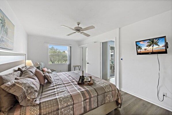 A bedroom with a bed, a ceiling fan, a TV on the wall, and a window with a view of trees.
