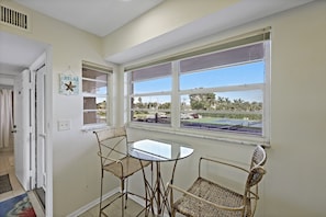 A room with a glass table and two wicker chairs, a window with a view of a parking lot, and a door with a towel rack. - Coastal Style Condo | Pool, Spa & Steps to Beach (Marco Island)