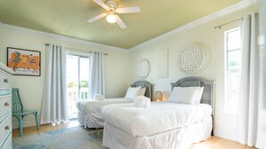 Une chambre avec deux lits, un ventilateur de plafond, une grande fenêtre avec des rideaux, une commode et un tapis.