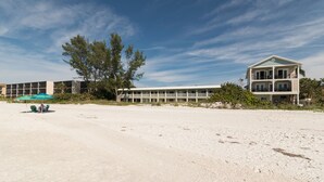 Un hotel frente a la playa con una estructura de dos pisos, una playa arenosa en primer plano y un cielo despejado.