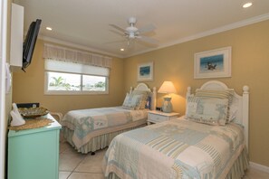 A bedroom with two beds, a ceiling fan, a window with blinds, and artwork on the walls. - Shell Cove 9 · Gulf Front · Beach Gear (Bradenton Beach)